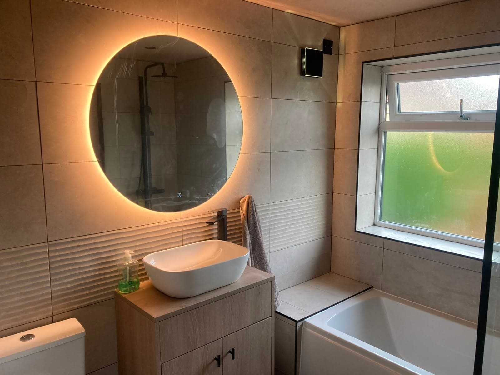 Warm neutral bathroom with a backlit circular mirror above an oak-effect vanity and countertop basin, beside a panelled bath