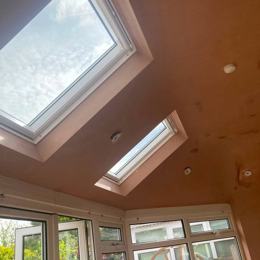 Inside a converted conservatory, looking up at a freshly plastered vaulted ceiling with two Velux roof windows and recessed downlights